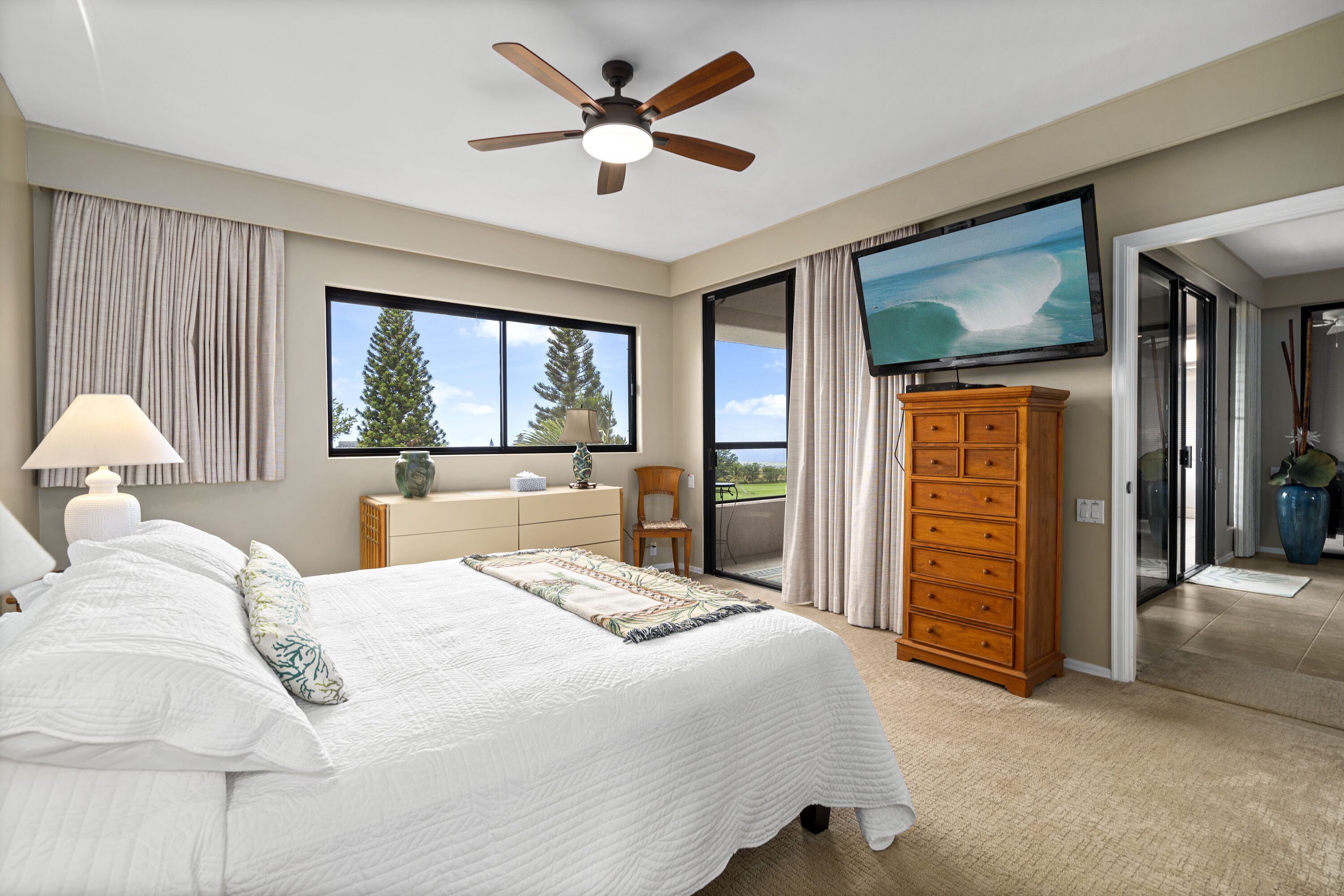A bedroom with a bed, a ceiling fan, a TV, a dresser, and a view of the outdoors.