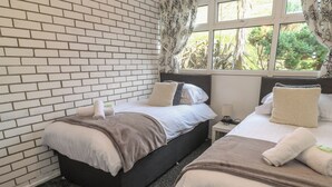 A hotel room with two beds, a brick-patterned wall, and a window with a view of greenery. - 16 Seaspray (Truro)