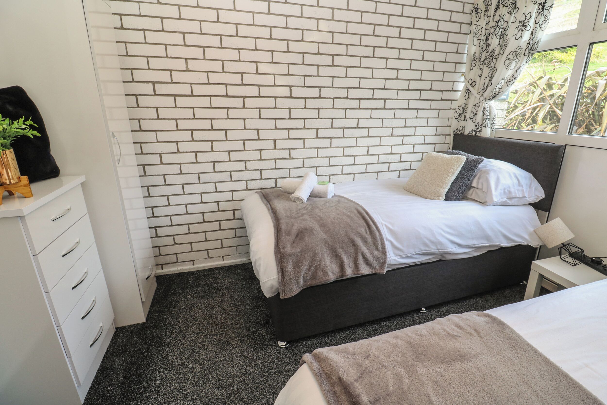 Ein Schlafzimmer mit Ziegelwand, einem Bett mit grauem Kopfteil, einem Nachttisch mit Lampe und einem Fenster mit Vorhängen.