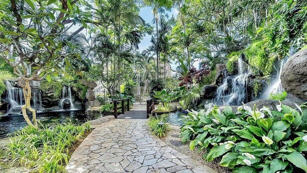 A stone pathway leading through a lush garden with multiple waterfalls and dense foliage.