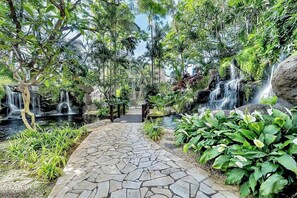 Um caminho de pedra serpenteia por um jardim exuberante, com várias cascatas e vegetação densa.