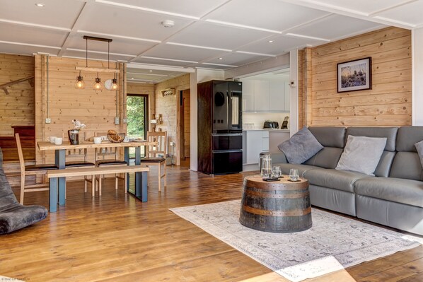 A modern living room with a grey sofa, wooden dining table, and a rustic barrel coffee table. - Buttercup Lodge | The Ridgeway Holiday Let | 2 Beds & Sleeps 4 (Benson)