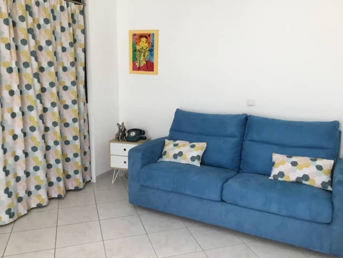 A room with a blue sofa, a white nightstand, a telephone, and a colorful painting on the wall.