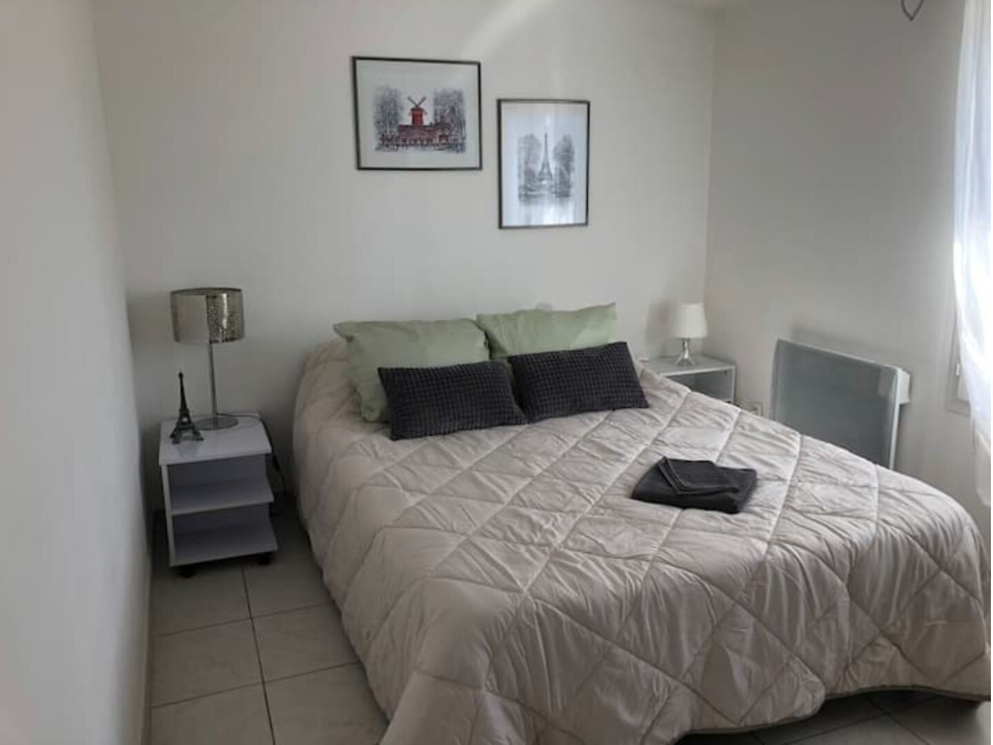 A neatly arranged bedroom with a bed, pillows, a bedside table, a lamp, and two framed pictures on the wall.