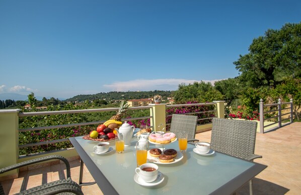 A breakfast table with fruits, pastries, and beverages on a balcony overlooking a scenic landscape.