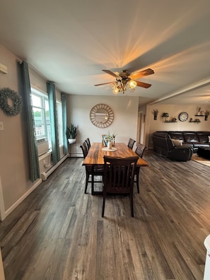 Uma sala de jantar com mesa e cadeiras de madeira, ventilador de teto, espelho fixado na parede, e uma sala de estar com sofá e relógio.