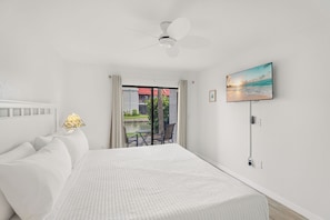 A bedroom with a bed, a ceiling fan, a TV, and a view of a garden through a glass door. - Spend a month away, just a quick walk to the sand! (Bradenton Beach)