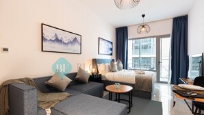 A modern living room with a grey sectional sofa, a small round table, and a bed with a view of the city. - Premium Studio in Oasis - 318 (Abu Dhabi)