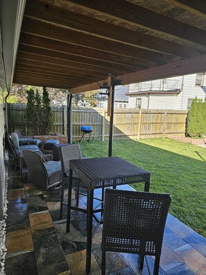 Ein überdachter Patio-Bereich mit Gartenmöbeln, einem Grill und Blick auf den Garten.