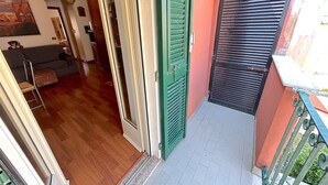 Ein schmaler Gang mit Holzboden führt zu einem Raum, der eine Couch und einen Sessel beherbergt. Eine grüne Tür und ein Fenster mit schwarzen Rollläden sind zu sehen.