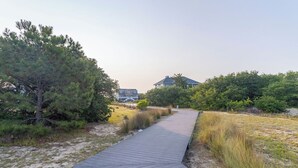 A wooden boardwalk leading to a house surrounded by dense greenery and a sandy area. - Near the Beach! Saltwater Pool, Hot Tub, Great Neighborhood, C0949 (Corolla)