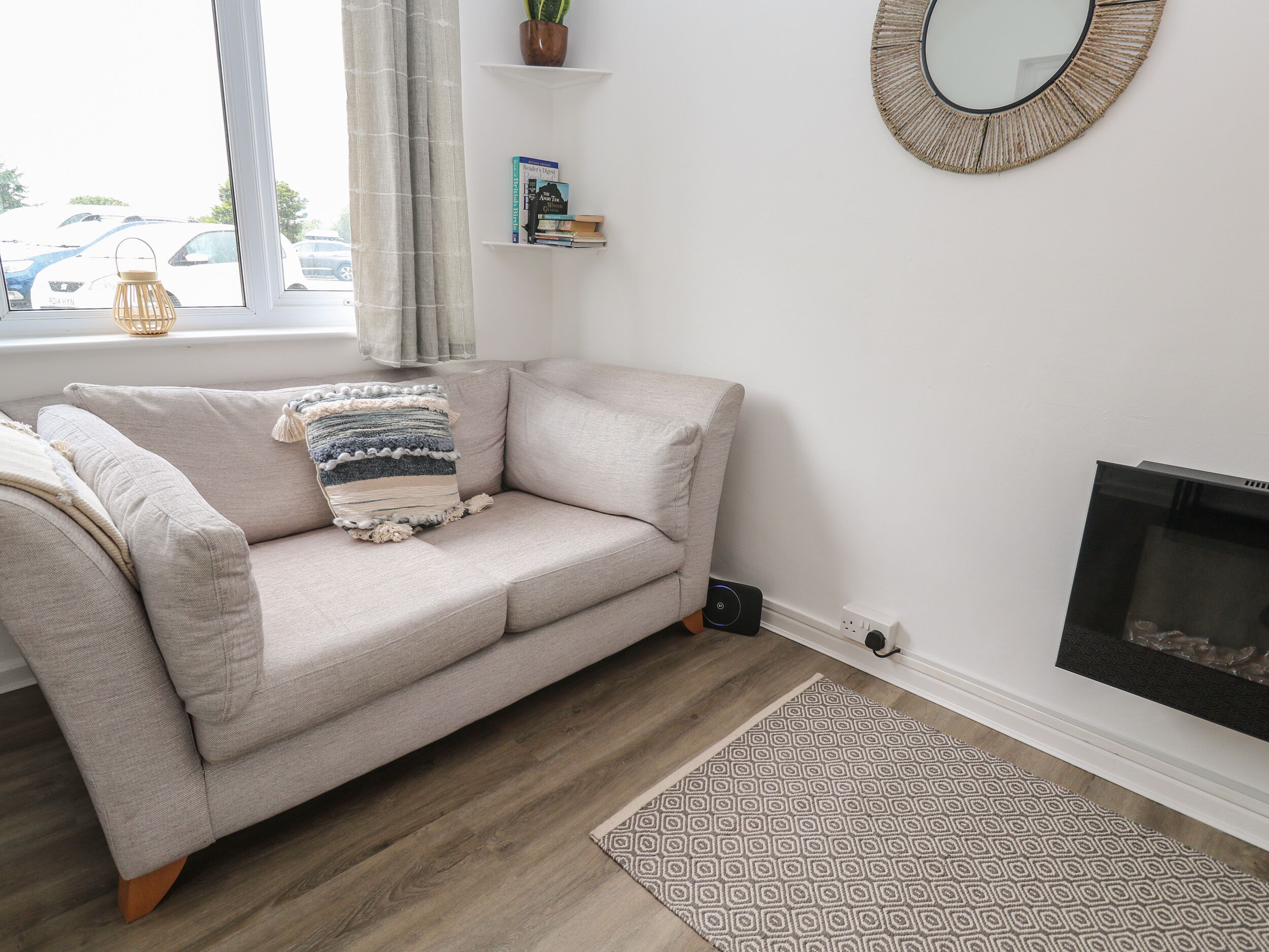 Una sala de estar con un sofá color beige, un cojín estampado, un estante con libros, un espejo redondo y una chimenea.