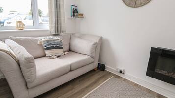 Una sala de estar con un sofá color beige, un cojín estampado, un estante con libros, un espejo redondo y una chimenea.