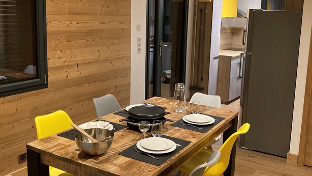 Un coin repas avec une table en bois, dressée pour quatre personnes et entourée de chaises, ainsi qu’une cuisine équipée d’un réfrigérateur et de placards.