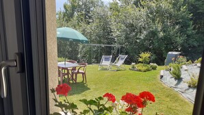 On aperçoit, à travers une porte vitrée, un jardin avec une pelouse, une table entourée de chaises, un parasol vert et un parterre de fleurs rouges.