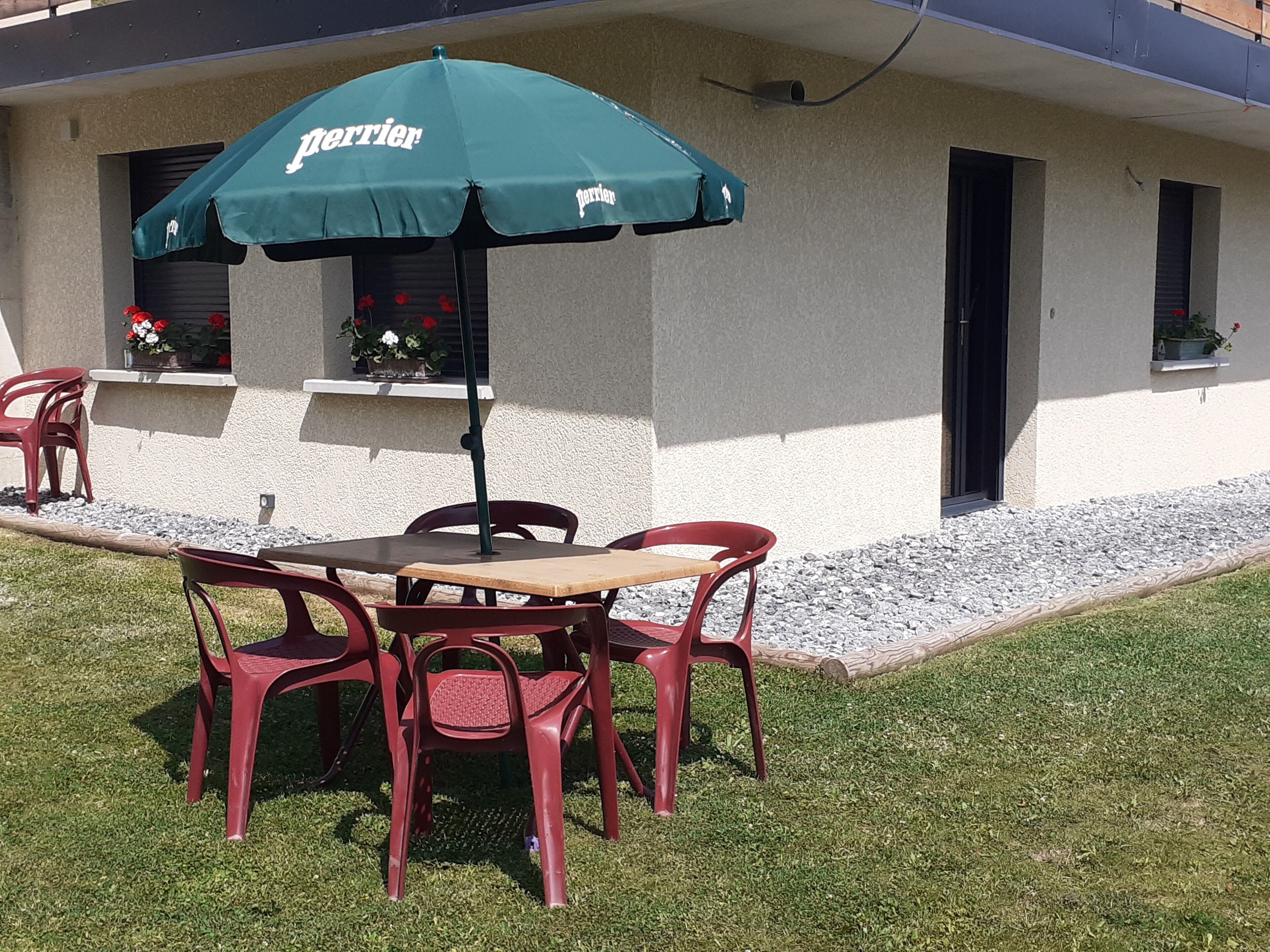 Un patio al aire libre con una mesa y cuatro sillas rojas, un paraguas verde con el logotipo 'Perrier' y un edificio de fachada blanca con ventanas adornadas con macetas de flores.