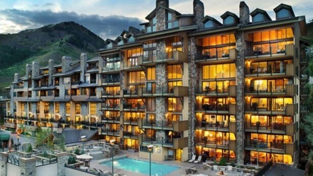 Un hotel de varios pisos con zona de piscina, mobiliario exterior y vistas a las montañas.