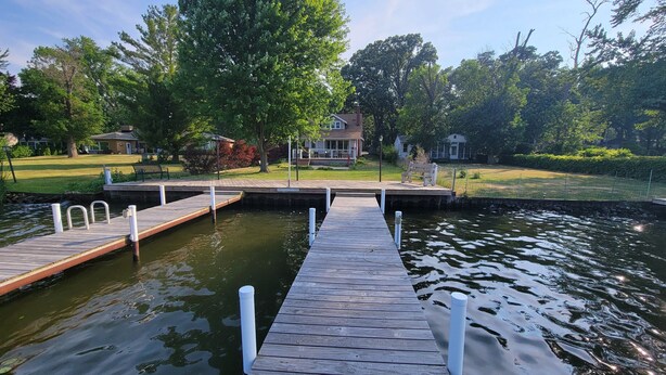 A wooden dock extending into a calm body of water, surrounded by lush greenery and a residential area with houses and a well-maintained lawn. - Riverfront Charmer with Character & Entertainment (McHenry)