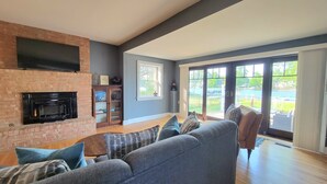 A living room with a fireplace, a sofa, and a view of a backyard through large sliding glass doors. - Riverfront Charmer with Character & Entertainment (McHenry)