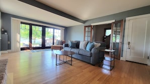 A spacious living room with a grey sofa, a coffee table, and a view of the outdoors through large windows. - Riverfront Charmer with Character & Entertainment (McHenry)