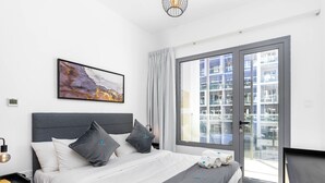 A modern hotel room with a large bed, a painting, a view of the city through a sliding glass door, and a small table with a lamp. - Modern Studio in Oasis -607 (Abu Dhabi)
