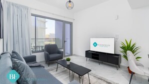A modern living room with a grey sofa, a black coffee table, a grey armchair, a TV on a black stand, and a balcony with a view of buildings. - Modern Studio in Oasis -607 (Abu Dhabi)