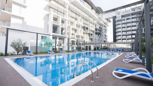 A modern hotel pool area with a clear blue pool, lounge chairs, and a multi-story building with balconies. - Modern Studio in Oasis -607 (Abu Dhabi)