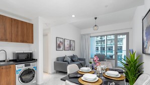 A modern apartment with a kitchen, dining area, and living space. It includes a washing machine, a microwave, a dining table set with plates and cutlery, and a sofa. - Modern Studio in Oasis -607 (Abu Dhabi)