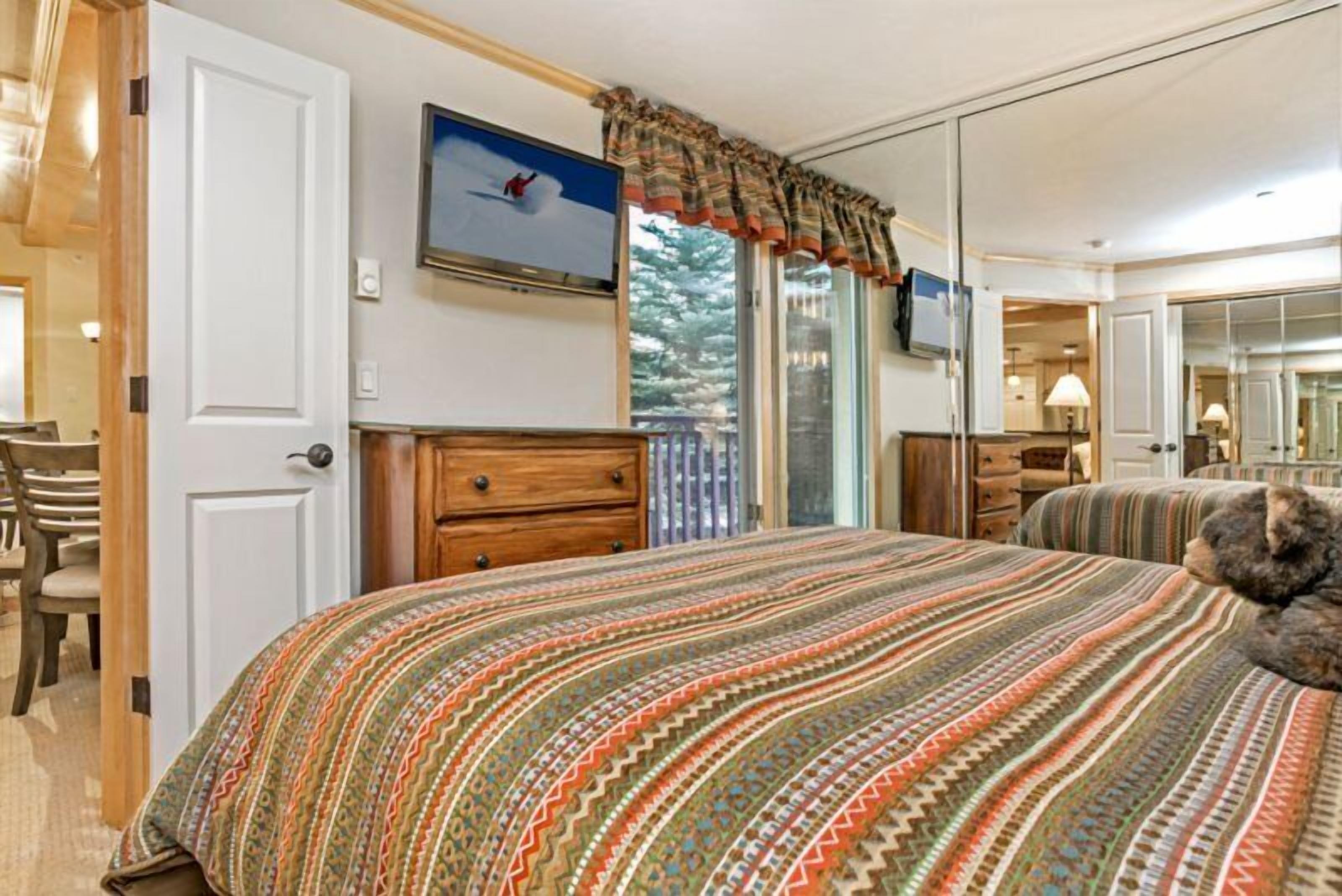 Een slaapkamer met een groot bed, een aan de muur bevestigde televisie en een raam met gordijnen.