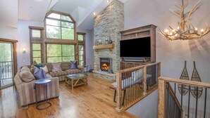 A spacious living room with a stone fireplace, a large TV, and a wooden staircase.