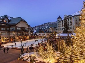 Ett snötäckt torg med en skridskobana, omgivet av byggnader med traditionell arkitektur och ett julgran i förgrunden.