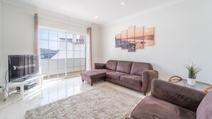A modern living room with a sectional sofa, a flat-screen TV, a large window with curtains, and a cityscape view. - Villa Lemos by HelloVacations (Albufeira)