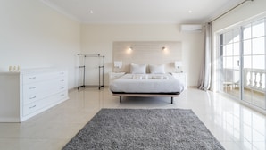 A modern bedroom with a large bed, a bedside table, a dresser, and a view of the outdoors through a large window. - Villa Lemos by HelloVacations (Albufeira)