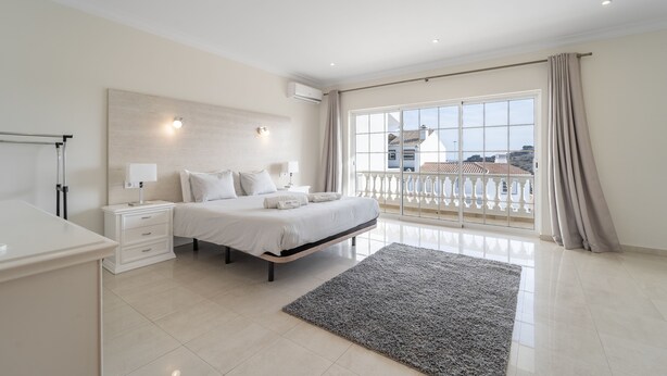 A modern bedroom with a large bed, bedside tables, a desk, and a balcony with a view. - Villa Lemos by HelloVacations (Albufeira)