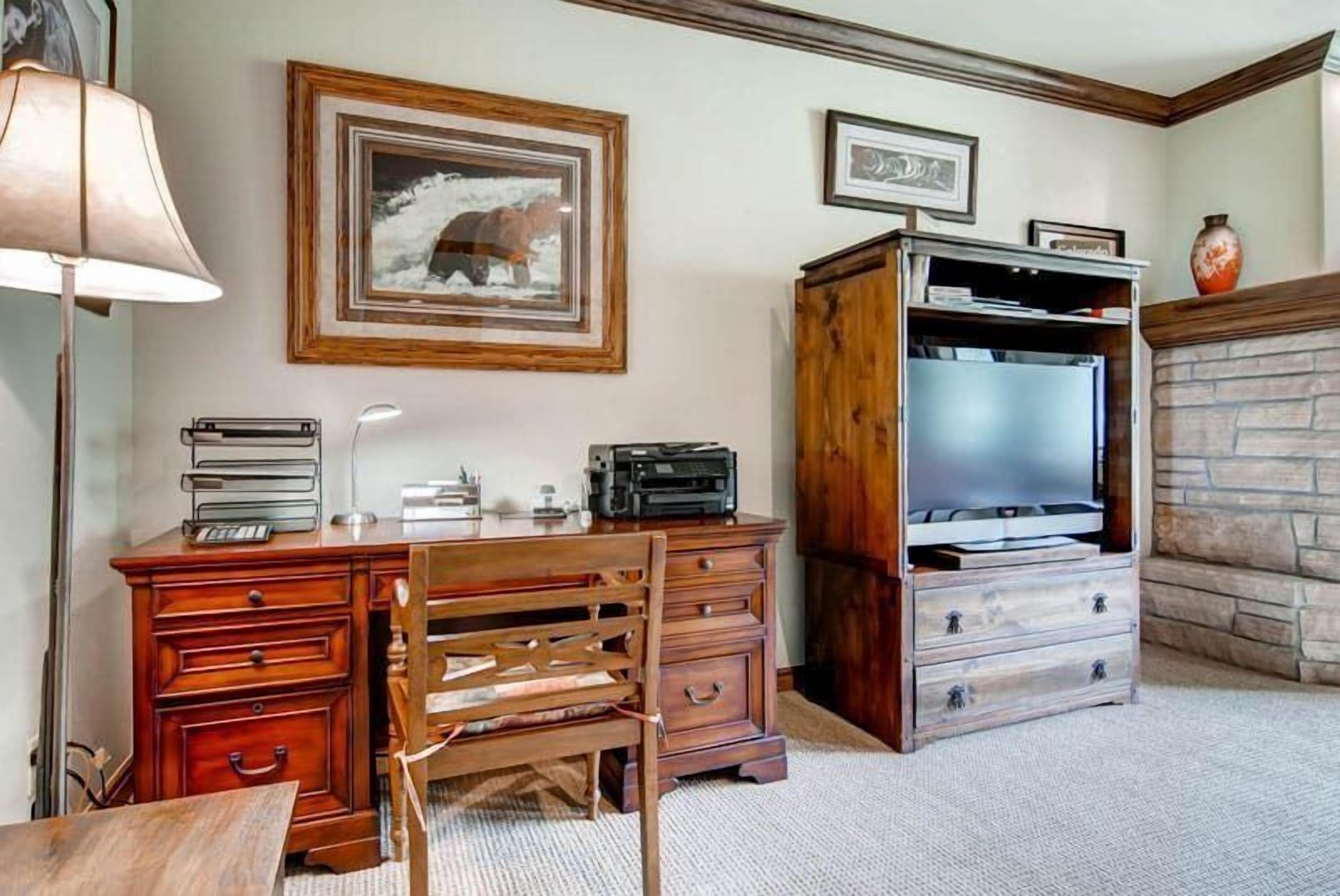 Une pièce avec un bureau en bois, une chaise, une lampe, un téléviseur posé sur un meuble en bois, des tableaux encadrés accrochés au mur et une cheminée en pierre.