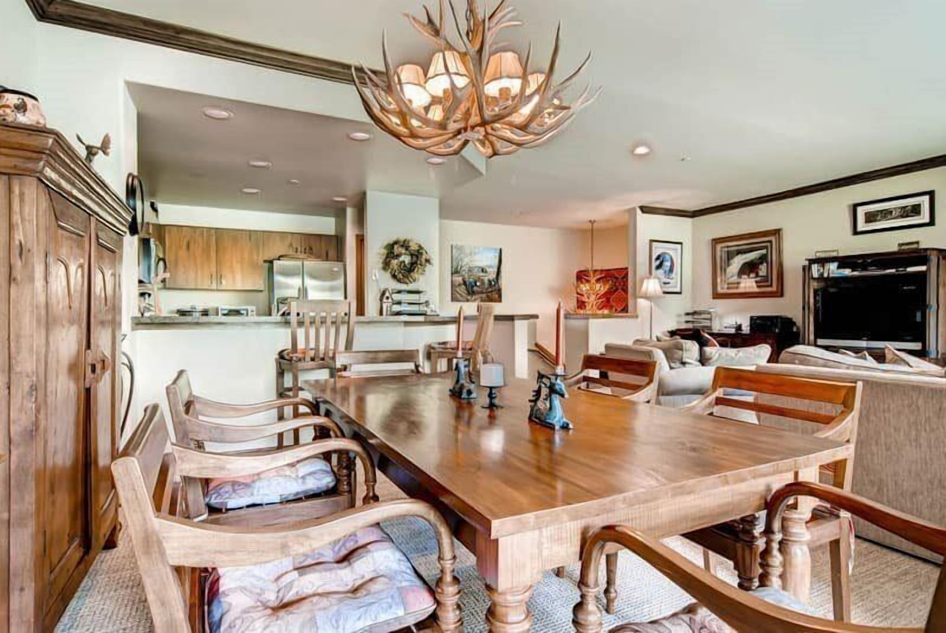 Un comedor amplio con una gran mesa de madera y sillas, una araña y una cocina al fondo.