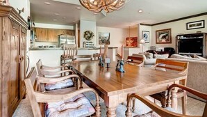 A spacious dining area with a large wooden table and chairs, a chandelier, and a kitchen in the background.