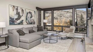 Un salon moderne avec un canapé d’angle gris, une table basse ronde et une vue sur un paysage montagneux grâce à de grandes fenêtres.
