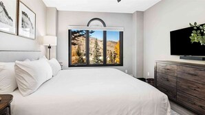 Une chambre avec un grand lit, une télévision, une commode et une fenêtre donnant sur des arbres et des montagnes.