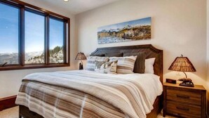 A bedroom with a large window, a bed with a striped blanket, two bedside lamps, and a painting of a mountain landscape.