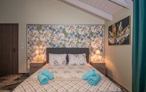 A bedroom with a checkered bedspread, floral pillows, two rolled towels, a headboard with a leaf pattern, bedside lamps, and a painting on the wall. - Casa Di Voz (Zakynthos)