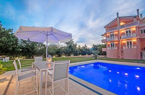 A poolside area with a table and chairs, a large umbrella, and a pink building with balconies.