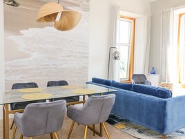 A modern living room with a glass dining table, grey upholstered chairs, a blue sofa, and a large abstract wall mural.