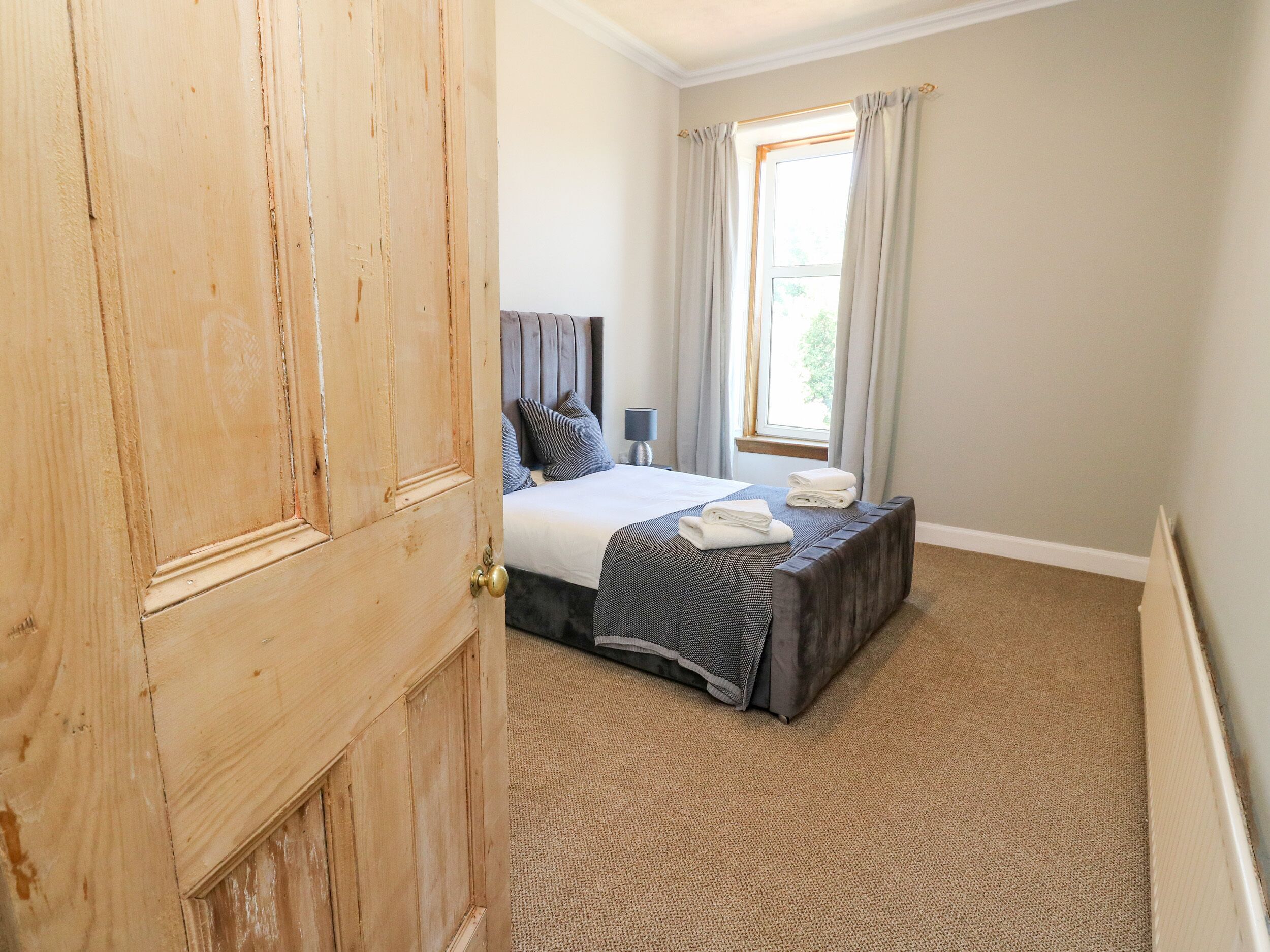 A bedroom with a bed, a nightstand, a window with curtains, and a wooden door.