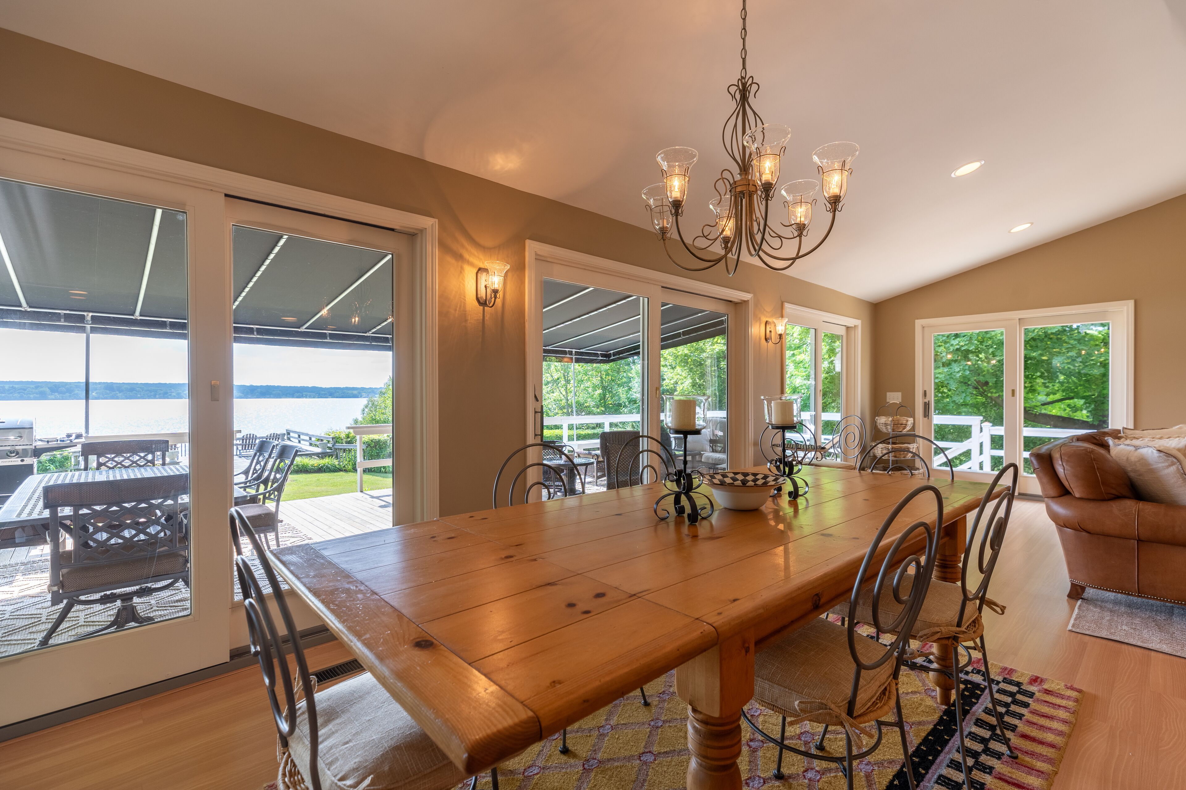 A spacious dining area with a large wooden table, metal chairs, and a chandelier. There are large windows and doors leading to an outdoor patio with a view of the water.