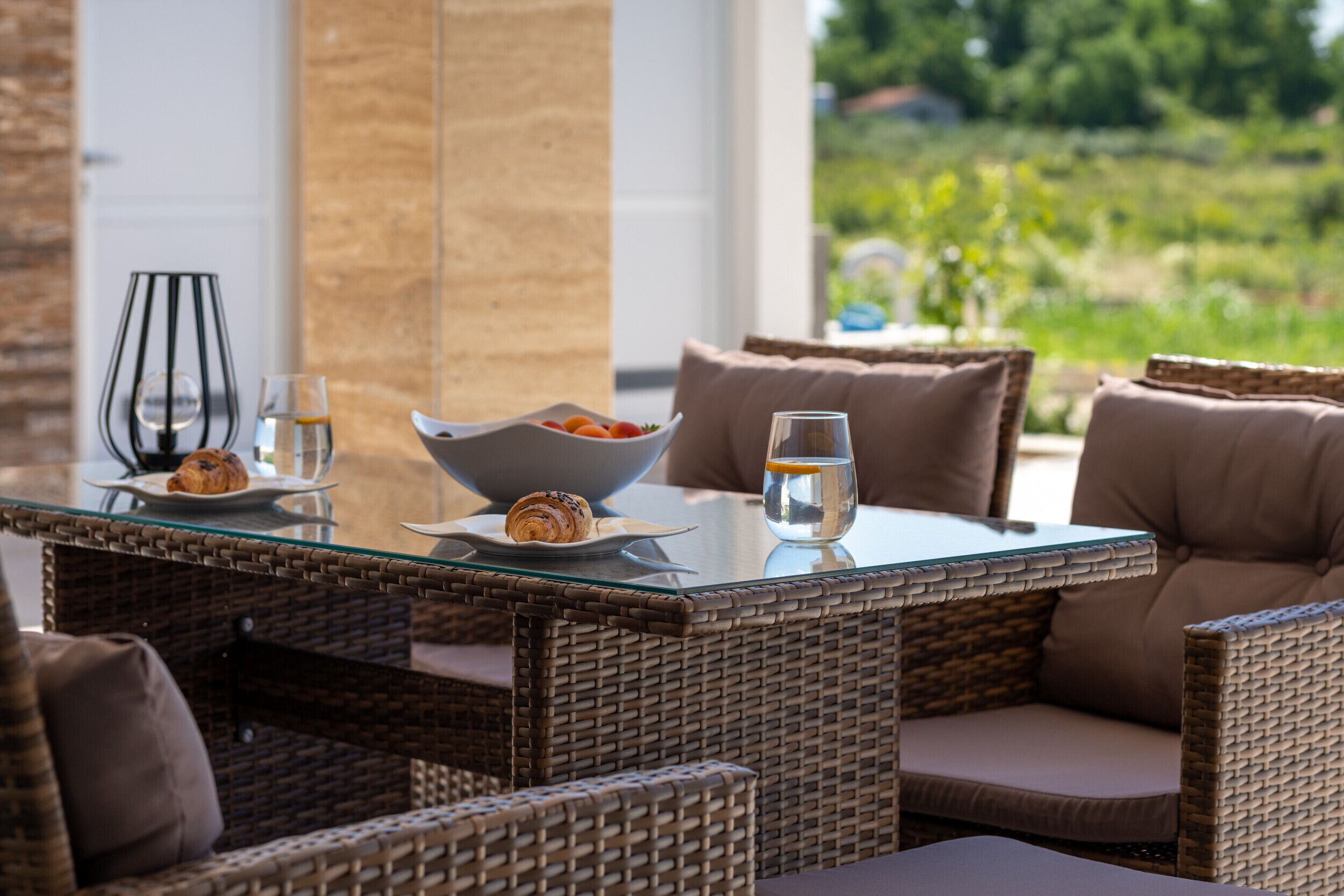 A wicker dining table with a glass top, surrounded by wicker chairs with cushions. A bowl of fruit and a croissant are on the table.