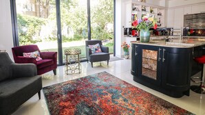 A modern kitchen with a large island, a dining area with a red sofa, and a view of greenery through large windows. - The Hideaway (Hove)