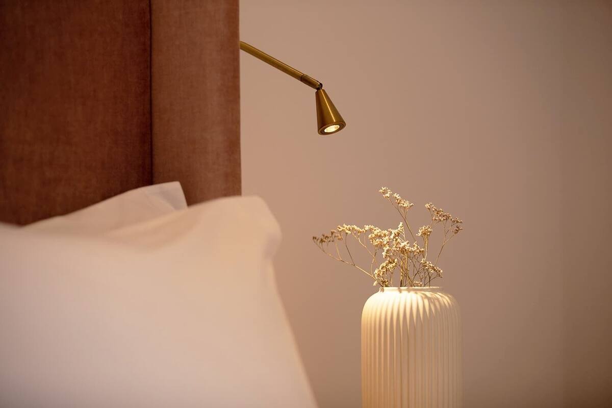 Un lit avec du linge de lit blanc, une table de chevet ornée d’un vase de fleurs séchées et une lampe fixée au mur.