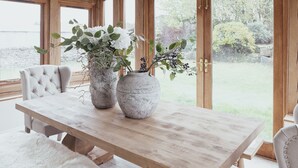 Una mesa de comedor de madera con dos grandes jarrones de piedra que contienen plantas, una silla con tapicería capitonada y vistas a un jardín a través de amplios ventanales de madera.