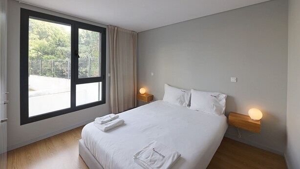 A hotel room with a large window, a bed with white linens, two bedside lamps, and a wooden nightstand. - FLH Boavista Flat I (Oporto)
